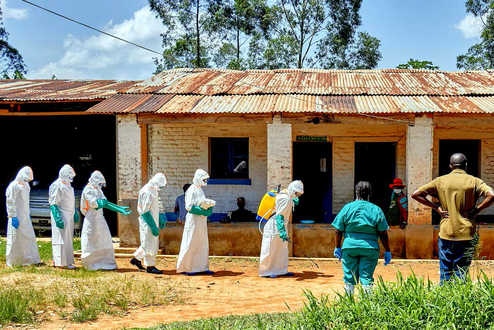 Trabajadores de la Cruz Roja, con equipo de protección personal (EPP), caminan en formación mientras uno de ellos desinfecta el camino en el hospital general de referencia de Mongbwalu (República Democrática del Congo), antes de manipular el cuerpo de un niño fallecido por ébola.