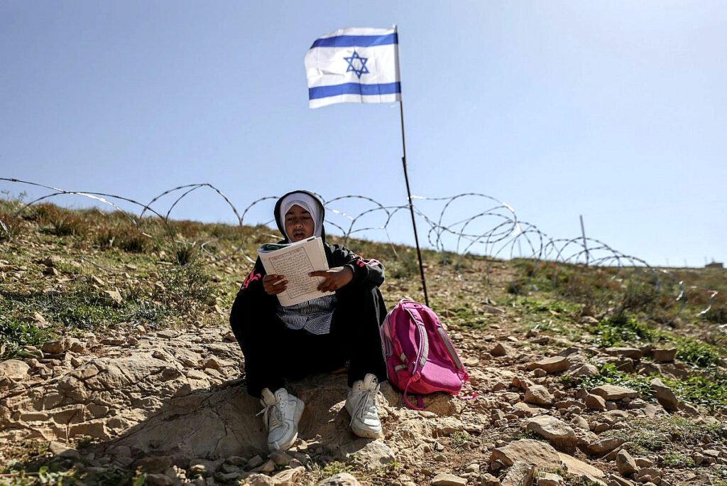 Una niña palestina lee su libro de texto frente a una valla de alambre de púas recién instalada con banderas israelíes, que los colonos israelíes colocaron para impedir el paso de profesores y alumnos palestinos desde sus hogares en la aldea de Umm Al-Khair hasta su escuela, Masafer Yatta, en la Cisjordania ocupada.