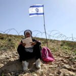 Una niña palestina lee su libro de texto frente a una valla de alambre de púas recién instalada con banderas israelíes, que los colonos israelíes colocaron para impedir el paso de profesores y alumnos palestinos desde sus hogares en la aldea de Umm Al-Khair hasta su escuela, Masafer Yatta, en la Cisjordania ocupada.