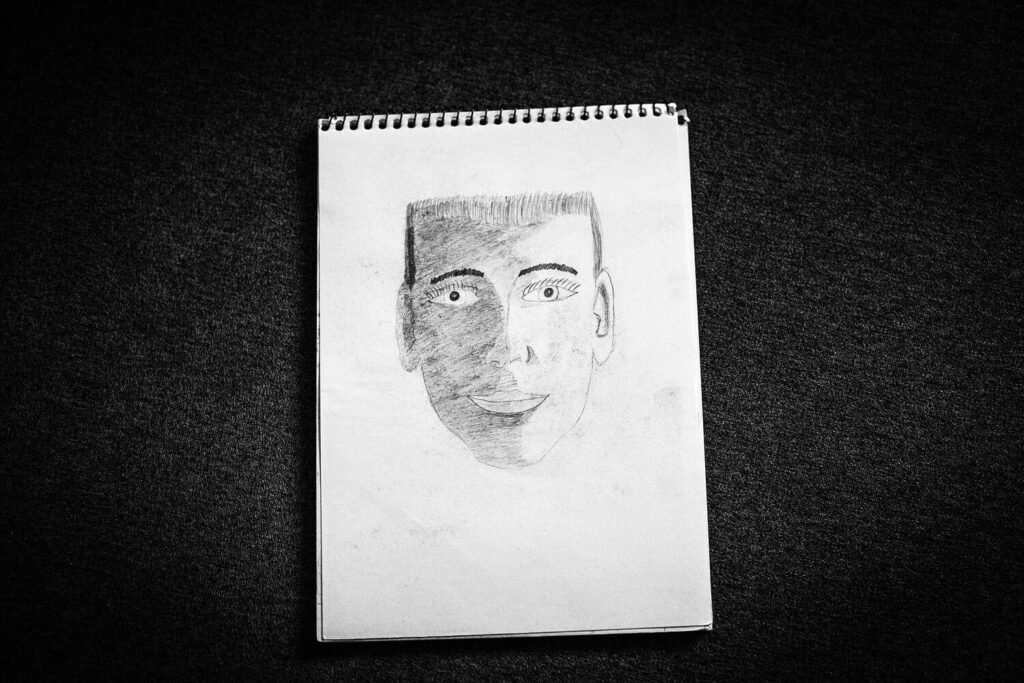 Un autorretrato de Alex muestra su rostro dividido entre luz y sombra, con los ojos abiertos en ambas mitades. La imagen revela la dualidad de su mundo interior y la complejidad de su presencia, dejando un testimonio íntimo de su experiencia.