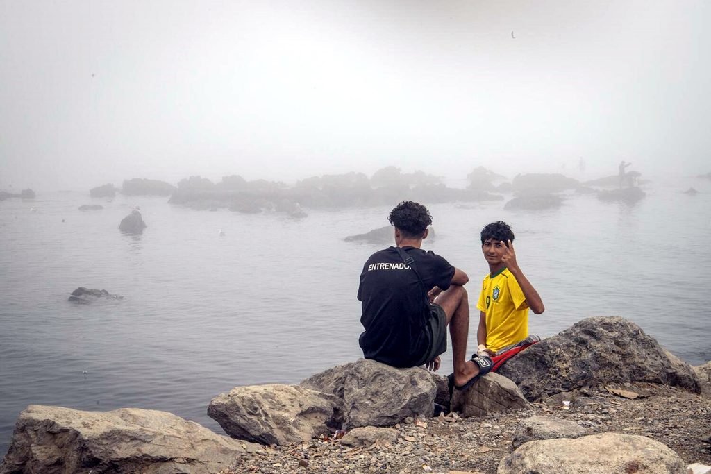 Un adulto y un menor en la playa del Trampolín sentados junto al mar.